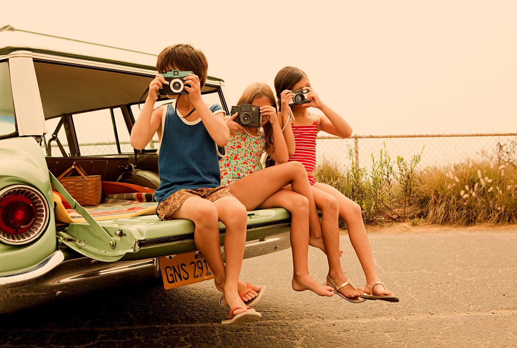 Three children sitting on the open tailgate of a vintage station wagon, each holding an old film camera up to one eye, a picnic basket and striped blanket behind them